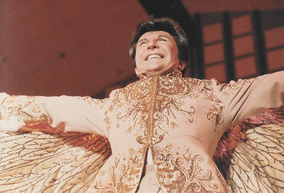 Ο Valentino Liberace [Βαλεντίνο Λιμπεράτσε] (1919–1987), Αμερικανός πιανίστας και διασκεδαστής, σε φωτογραφία του Alan Light (1983). Με τα εξεζητημένα κοστούμια, τη θεατρική περσόνα και τη μετατροπή του πιανιστικού ρεσιτάλ σε πολυτελές θέαμα, ο Λιμπεράτσε καθιερώθηκε ως μία από τις χαρακτηριστικότερες μορφές του μουσικού σόου μαν του 20ού αιώνα.[Φωτογραφία: Alan Light, 1983, CC BY 2.0, μέσω Wikimedia Commons]