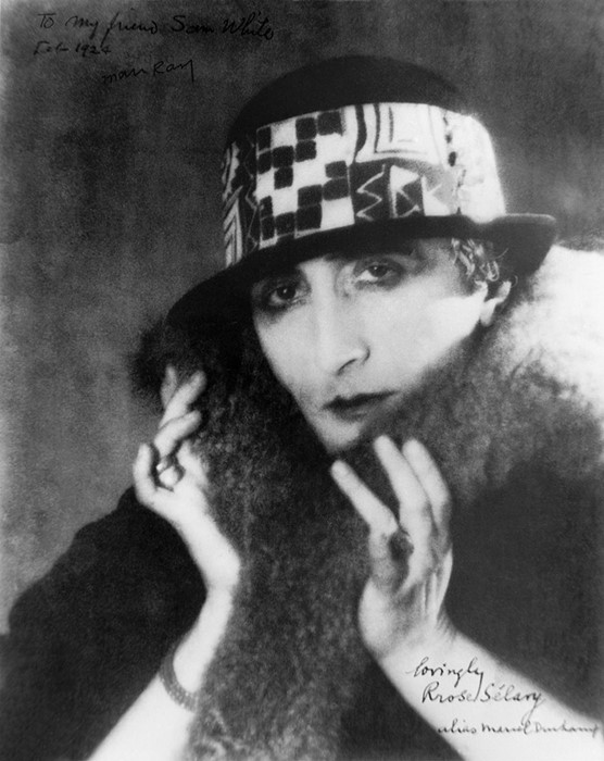 Marcel Duchamp as Rrose Sélavy [Ο Μαρσέλ Ντυσάν ως Ρρόζ Σελαβύ], φωτογραφία του Man Ray [Μαν Ραίη] (περ. 1920-1921). Με γούνινο κολάρο, καπέλο και ψιμυθίωση, ο Marcel Duchamp [Μαρσέλ Ντυσάν] υποδύεται τη θηλυκή περσόνα του, που το όνομά της κρύβει το λογοπαίγνιο «Éros, c'est la vie» [«Έρως, αυτή είναι η ζωή»]. Η Rrose Sélavy υπέγραφε έργα και κείμενα, οπότε το κοινωνικό φύλο του δημιουργού γίνεται κι αυτό επινόηση: μια υπογραφή που μπορεί να αλλάξει.