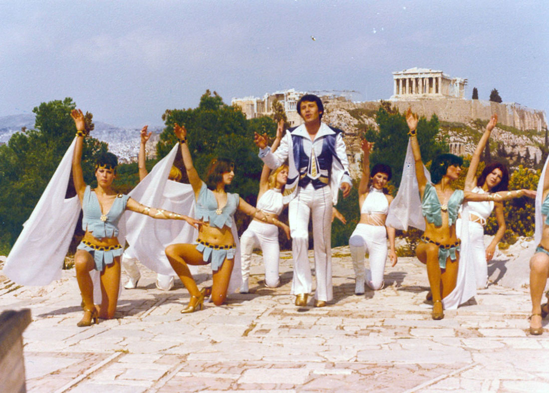 Ο Γιάννης Φλωρινιώτης ως ο λαϊκός σταρ Ντίμης Φλεριανός, πλαισιωμένος από χορεύτριες μπροστά στην Ακρόπολη, στην ταινία Και ξανά προς τη δόξα τραβά (1980) του [[#Γιώργου Σταμπουλόπουλου#]]. Η εικόνα συμπυκνώνει, με ειρωνική και παρωδιακή διάθεση, την ελληνοπρέπεια, τη φαντασμαγορία και την αισθητική των σόου πίστας, που η ταινία αξιοποιεί για να σατιρίσει τη μαζική ψυχαγωγία και την κατασκευή του λαϊκού ειδώλου. 