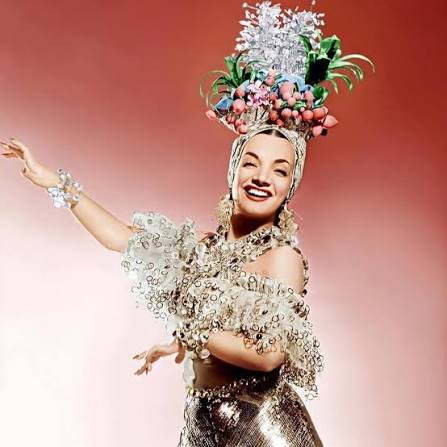 Η [[#Carmen Miranda [Κάρμεν Μιράντα]#]], φωτογραφία δημοσιότητας, δεκαετία του 1940. Το κάλυμμα κεφαλής με φρούτα και άνθη, τα μεταλλικά κοσμήματα και η στυλιζαρισμένη χειρονομία συνθέτουν την περσόνα που η αμερικανική βιομηχανία του θεάματος κατασκεύασε γύρω από την Βραζιλιάνα ερμηνεύτρια της σάμπας. Η υπερβολή του κοστουμιού και η εμπορευματοποιημένη «εξωτική» θηλυκότητα κατέστησαν τη Miranda πρότυπο του αθέλητου καμπ και σταθερή αναφορά της ντραγκ εικονογραφίας.