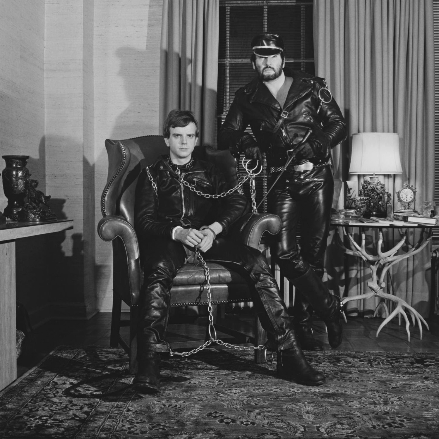 Robert Mapplethorpe [Ρόμπερτ Μάπλθορπ], Brian Ridley and Lyle Heeter, 1979. Το ζεύγος φωτογραφίζεται με δερμάτινα, αλυσίδες και μαστίγιο σε αστικό σαλόνι, ο υποτασσόμενος δεμένος σε πολυθρόνα και ο κυρίαρχος όρθιος δίπλα του. Η αντίστιξη σαδομαζοχιστικής σκευής και μικροαστικού διακόσμου φέρνει τις πρακτικές σε διάλογο με την κουήρ θεωρία και το κοινωνικό φύλο.