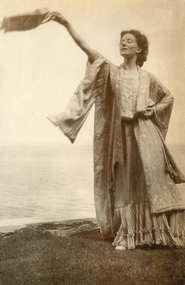 Εύα Πάλμερ Σικελιανού [Eva Palmer-Sikelianou] σε υπαίθρια χορευτική στάση, με ασάνδαλα πόδια και χειροϋφασμένο μεταξωτό ένδυμα. Η λήψη συνοψίζει τις αρχές που τροφοδότησαν τον μοντέρνο χορό: απελευθέρωση του σώματος από τον κορσέ και το χορευτικό υπόδημα, χειρονομία που εκτείνεται στο φυσικό τοπίο, αναγωγή στο αρχαιοελληνικό ιδεώδες ως εναλλακτική στη γεωμετρική πειθαρχία του κλασικού μπαλέτου.
