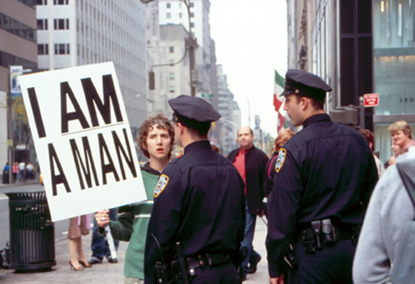 Η [[#Sharon Hayes [Σάρον Χέυς]#]] με το πλακάτ «I AM A MAN» [«Είμαι άνθρωπος / Είμαι άνδρας»] στη Νέα Υόρκη, στο In the Near Future [Στο εγγύς μέλλον] (2005). Το σύνθημα των απεργών εργατών καθαριότητας του Μέμφις του 1968 διεκδικούσε ανθρώπινη αξιοπρέπεια. Στα χέρια μιας γυναίκας το 2005 διαβάζεται ως δήλωση φύλου. Η επιτελεστικότητα του εκφωνήματος εξαρτάται από το πλαίσιο: η ίδια φράση, όταν επαναλαμβάνεται αλλού, γίνεται άλλη πράξη.