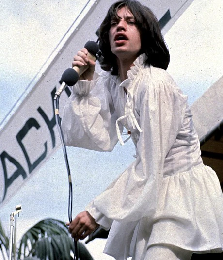 Ο Mick Jagger [Μικ Τζάγκερ] στην ιστορική συναυλία των Rolling Stones στο Χάιντ Παρκ το 1969. Ανατρέποντας τα έμφυλα πρότυπα της εποχής με το χαρακτηριστικό λευκό χιτώνιο και εκπέμποντας μια αχαλίνωτη διονυσιακή ενέργεια, ο καλλιτέχνης κυριαρχεί στο πλήθος με την αύρα ενός σχεδόν θρησκευτικού ηγέτη. Η επιβλητική, γεμάτη ερωτισμό και αλαζονεία σκηνική του παρουσία τον καθιερώνει ως το απόλυτο «ιερό τέρας» της ροκ κουλτούρας.