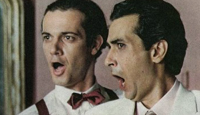 Ο Γιώργος Κιμούλης και ο Κώστας Αρζόγλου στην παράσταση Ζητείται τενόρος [Lend Me a Tenor, 1986] του Ken Ludwig [Κεν Λούντβιχ], σε σκηνοθεσία Κώστα Αρζόγλου, με σκηνικά και κοστούμια Γιώργου Ασημακόπουλου, στο Θέατρο Σμαρούλα (Αθήνα, σαιζόν 1989–1990). Δημοφιλής αμερικανική φάρσα που μετέφερε τις τεχνικές των μεταμφιέσεων, των ψευδών ταυτοτήτων και του φρενιτιώδους ρυθμού στον κόσμο της οπερατικής σκηνής του Μεσοπολέμου. 