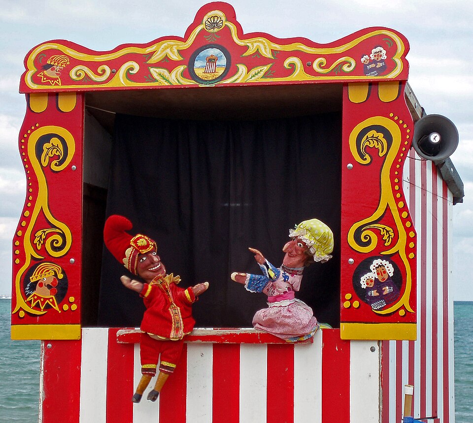 Παραδοσιακό θεατράκι Punch and Judy [Παντς και Τζούντυ] στο Swanage [Σουόνατζ] του Dorset [Ντόρσετ] της Αγγλίας. Το βρετανικό αυτό είδος κουκλοθεάτρου, με ρίζες στην ιταλική κομμέντια ντελλ άρτε και τη μορφή του Pulcinella [Πουλτσινέλλα], στήνεται σε όρθια, στενή παράγκα, όπου ο/η κουκλοπαίκτης/κουκλοπαίκτρια εμψυχοποιεί τις γαντόκουκλες πάνω από το κεφάλι του/της. Φωτογραφία: ALoan, 2006. Wikimedia Commons.