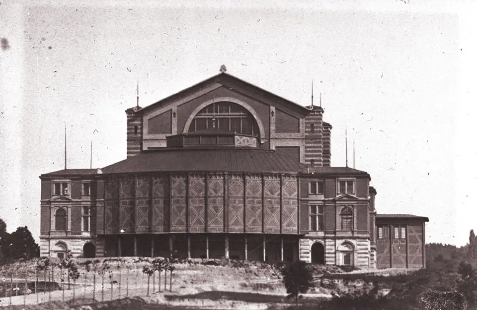 Ιστορική φωτογραφία του Bayreuther Festspielhaus [Φεστιβαλικού Θεάτρου του Μπαϋρόυτ] πριν από το 1882, χωρίς την προσθήκη του βασιλικού οικήματος [Königsbau]. Σχεδιασμένο από τον Richard Wagner σε συνεργασία με τον αρχιτέκτονα Otto Brückwald [Όττο Μπρύκβαλντ] και εγκαινιασμένο το 1876 με την πρώτη ολοκληρωμένη παράσταση του Der Ring des Nibelungen [Το Δαχτυλίδι των Νιμπελούνγκεν], το θέατρο συνιστά την αρχιτεκτονική και θεσμική υλοποίηση της βαγκνερικής σύλληψης του ολοτεχνήματος. © Richard-Wagner-Archiv, Nationalarchiv Bayreuth.