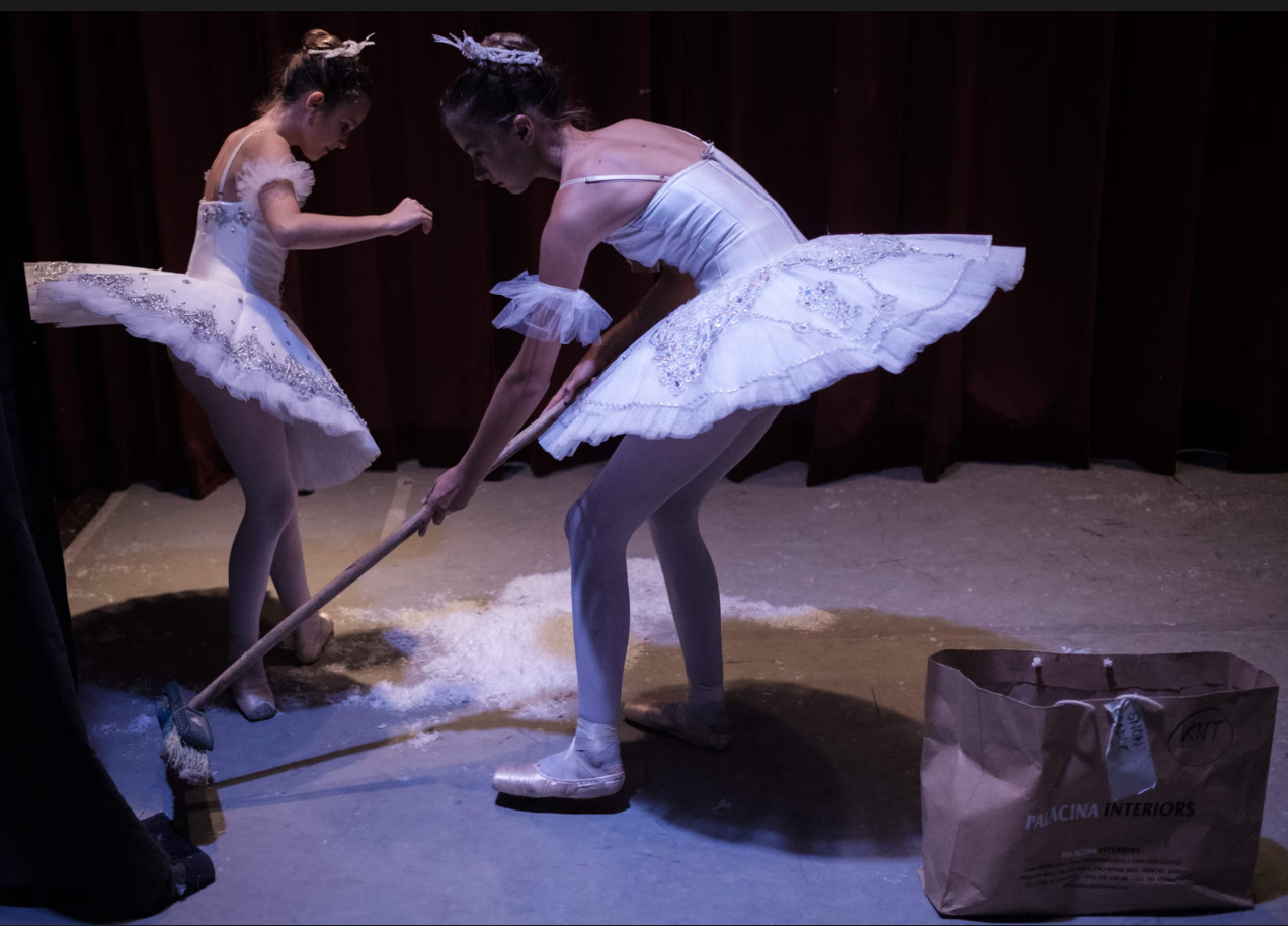 Δύο νεαρές χορεύτριες του Dance Centre Kenya καθαρίζουν τις τεχνητές νιφάδες χιονιού από τη σκηνή κατά το διάλειμμα παράστασης του Καρυοθραύστη [The Nutcracker]. Φορώντας ακόμη τα λευκά τους τουτού, αναλαμβάνουν εργασίες σκηνικής συντήρησης που κανονικά παραμένουν αόρατες, αποκαλύπτοντας το διάλειμμα ως χρόνο πρακτικής, παρασκηνιακής μέριμνας πίσω από τη μαγεία του θεάματος. Φωτογραφία: Yasuyoshi Chiba / AFP / Getty Images.