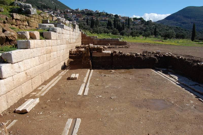 Οι λίθινες αύλακες υποδοχής των τροχών της κινητής σκηνής κατά μήκος της ανατολικής παρόδου του αρχαίου θεάτρου της Μεσσήνης. 